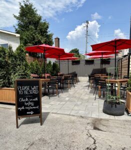 Image of a restaurant patio. Six wooden squared patio tables with six red patio umbrellas and four black chairs at each table. The sign at the entrance says, "Please wait to be seated. Check in with the hostess inside the restaurant."