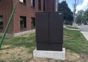 A tall slim utility box on a concrete base at the corner of two roads