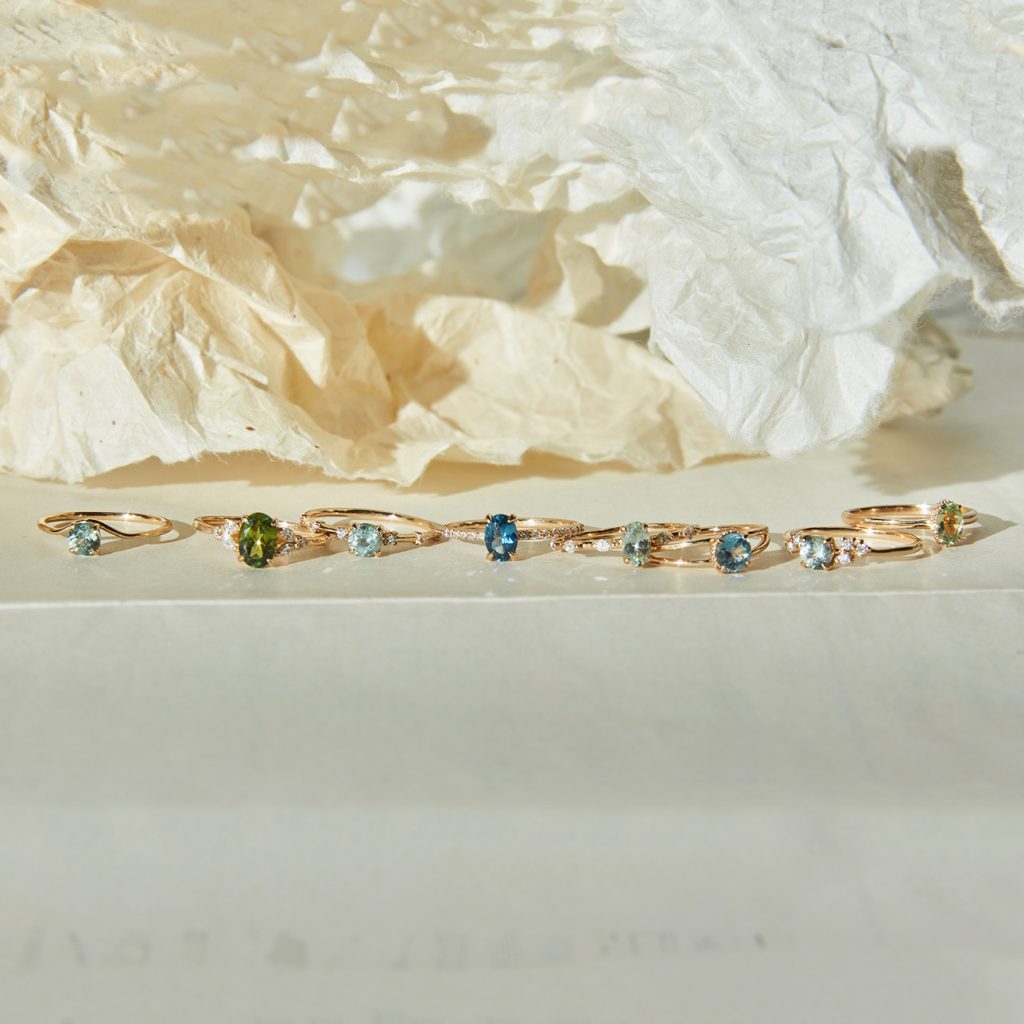 Eight gold rings with different coloured semi-precious stones on a white stone surfance