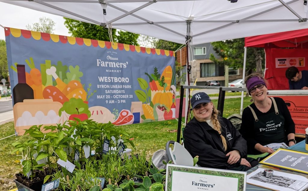 Two women sitting at a table with garden plants in pots and a banner behind with written details for the farmers market