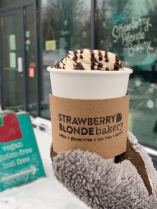 A paper to go cup with whip cream and chocolate sauce being held by a right hand with a grey mitten on