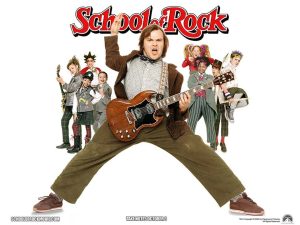 A man in brown pants, beige shirt, bowtie and blazer standing wide legged with a guitar, and children standing behind